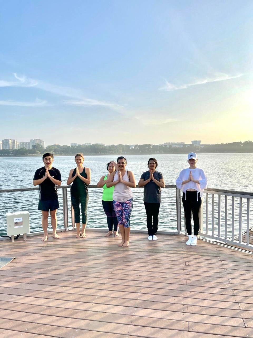 A YOGAM group standing in prayer pose at sunrise on a lakeside deck
